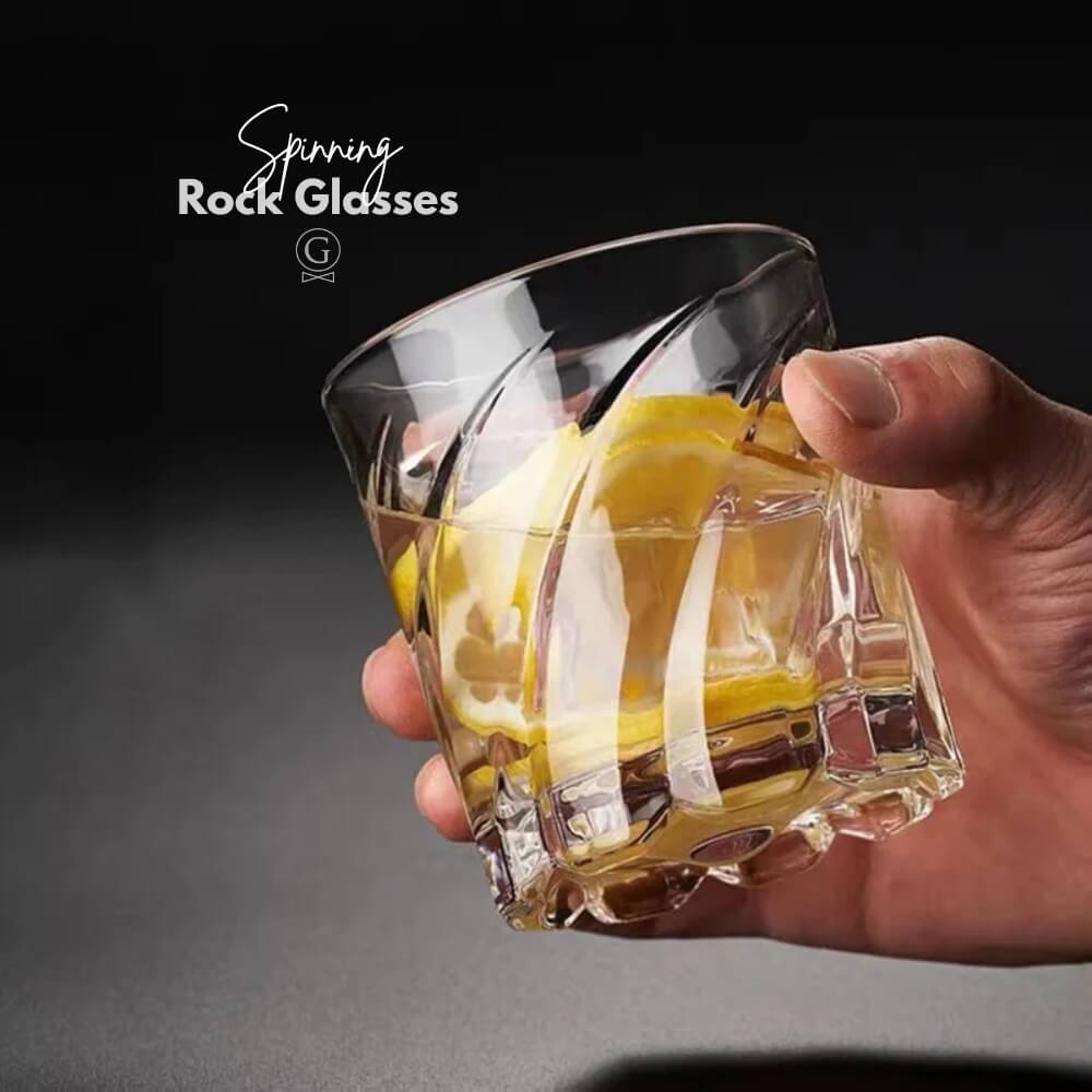 Hand holding a glass with a lemon slice against a dark background, featuring the brand 'Spinning Rock Glasses'.